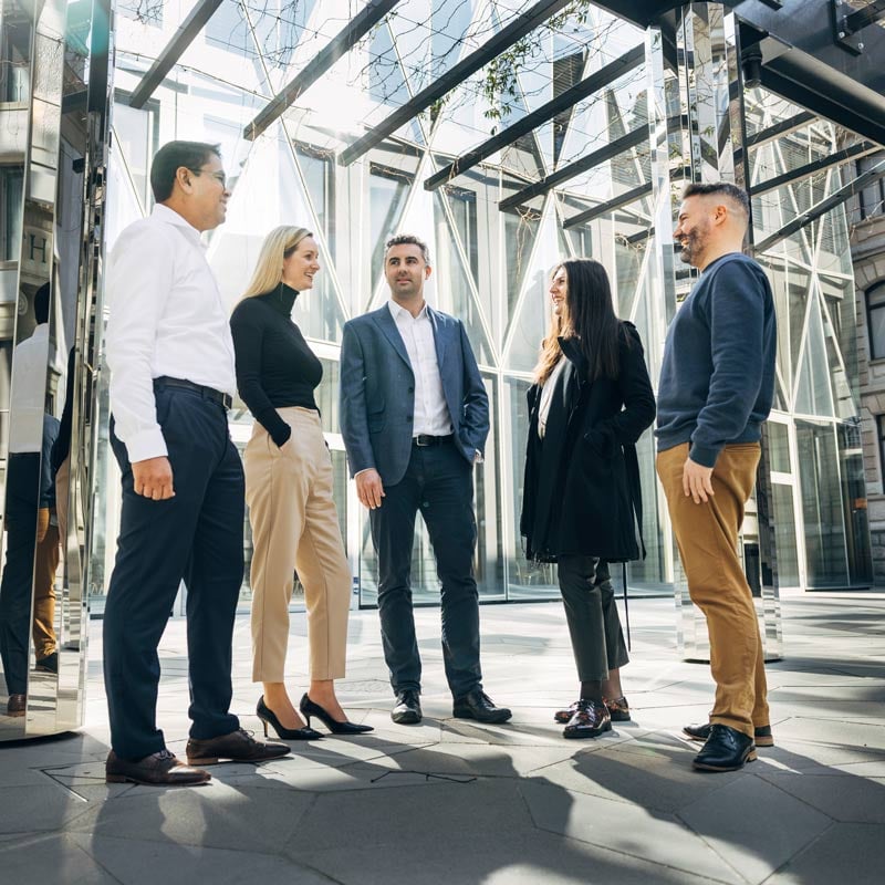 Eine Gruppe an fünf Menschen, drei Männer und zwei Frauen, stehen im Businessoutfit vor einem imposanten Firmengebäude. 